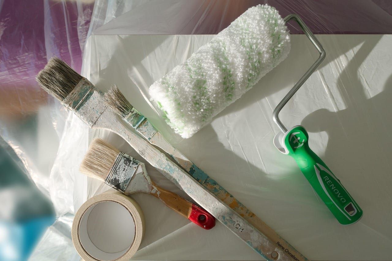 Ferramentas de pintura incluindo rolo, pincéis e fita crepe sobre uma superfície clara e plástico protetor.